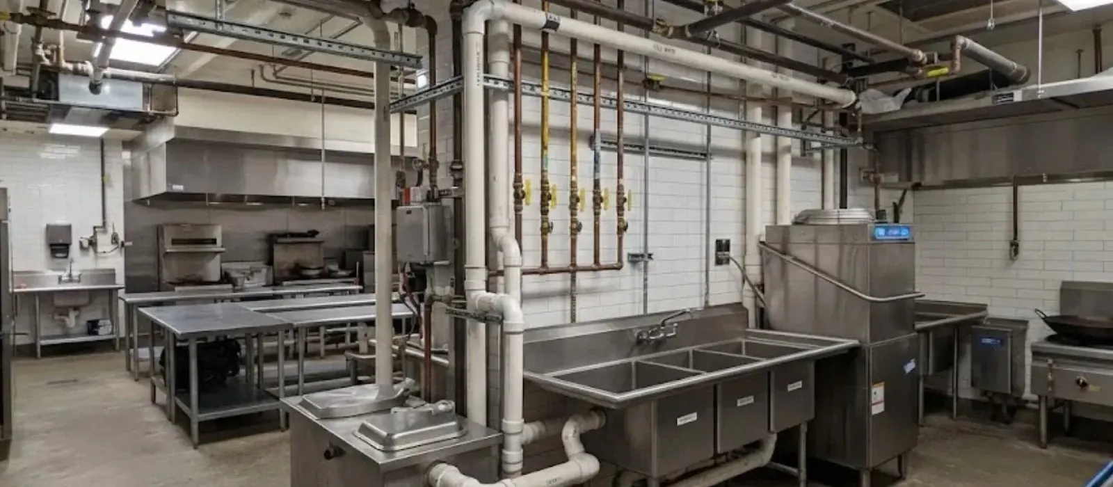 A commercial kitchen with exposed overhead pipes and a stainless steel sink and grease interceptor
