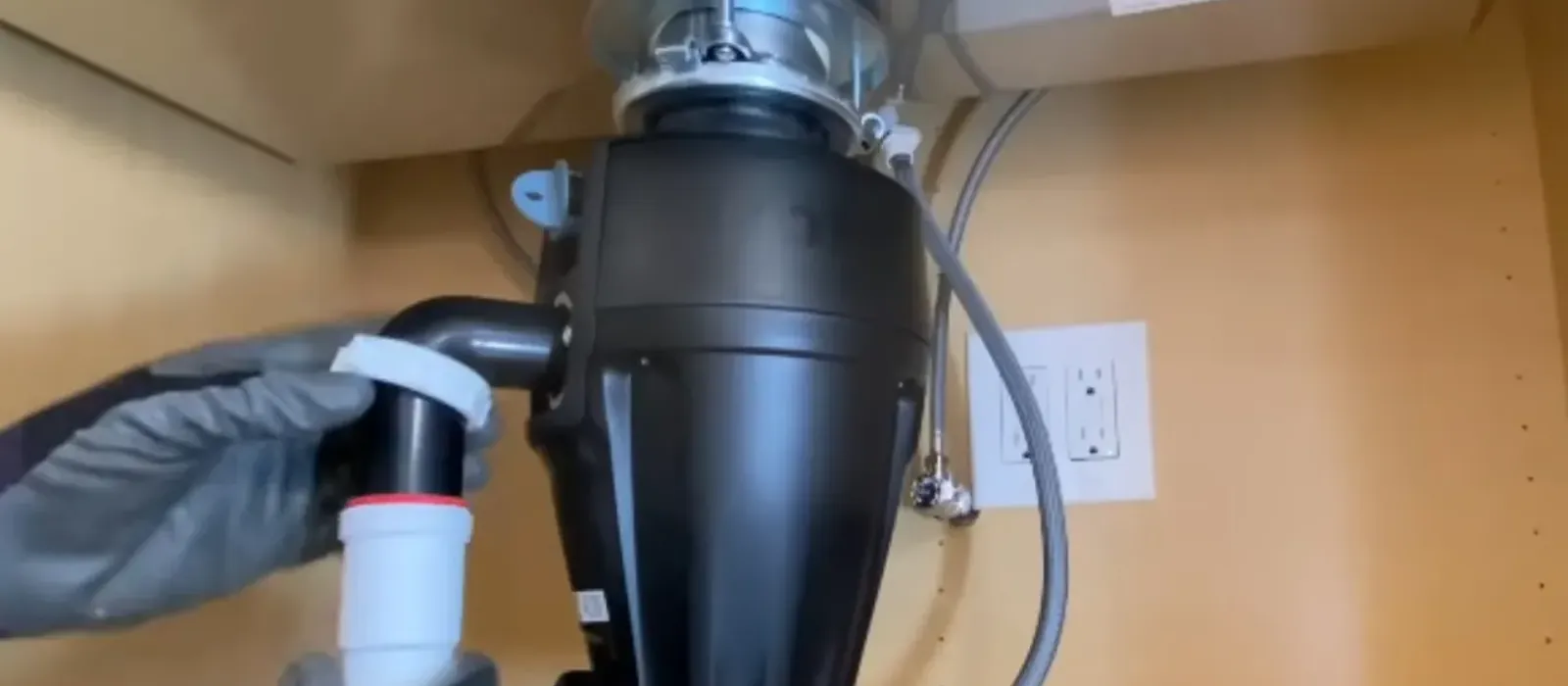 Gloved hands connecting the drain pipe to a garbage disposal mounted under a kitchen sink